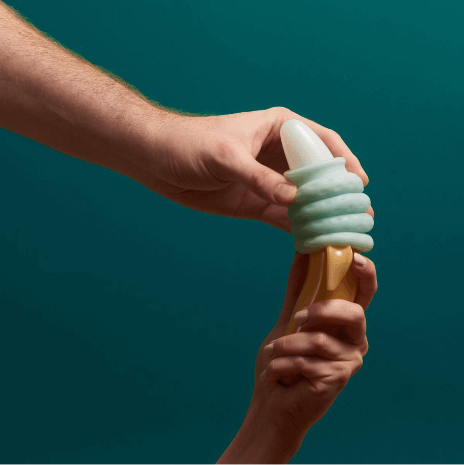 One hand holds a glass banana over a teal background. Another hand squishes the Ohnut rings down on the banana—showing how Ohnut compresses during sex.